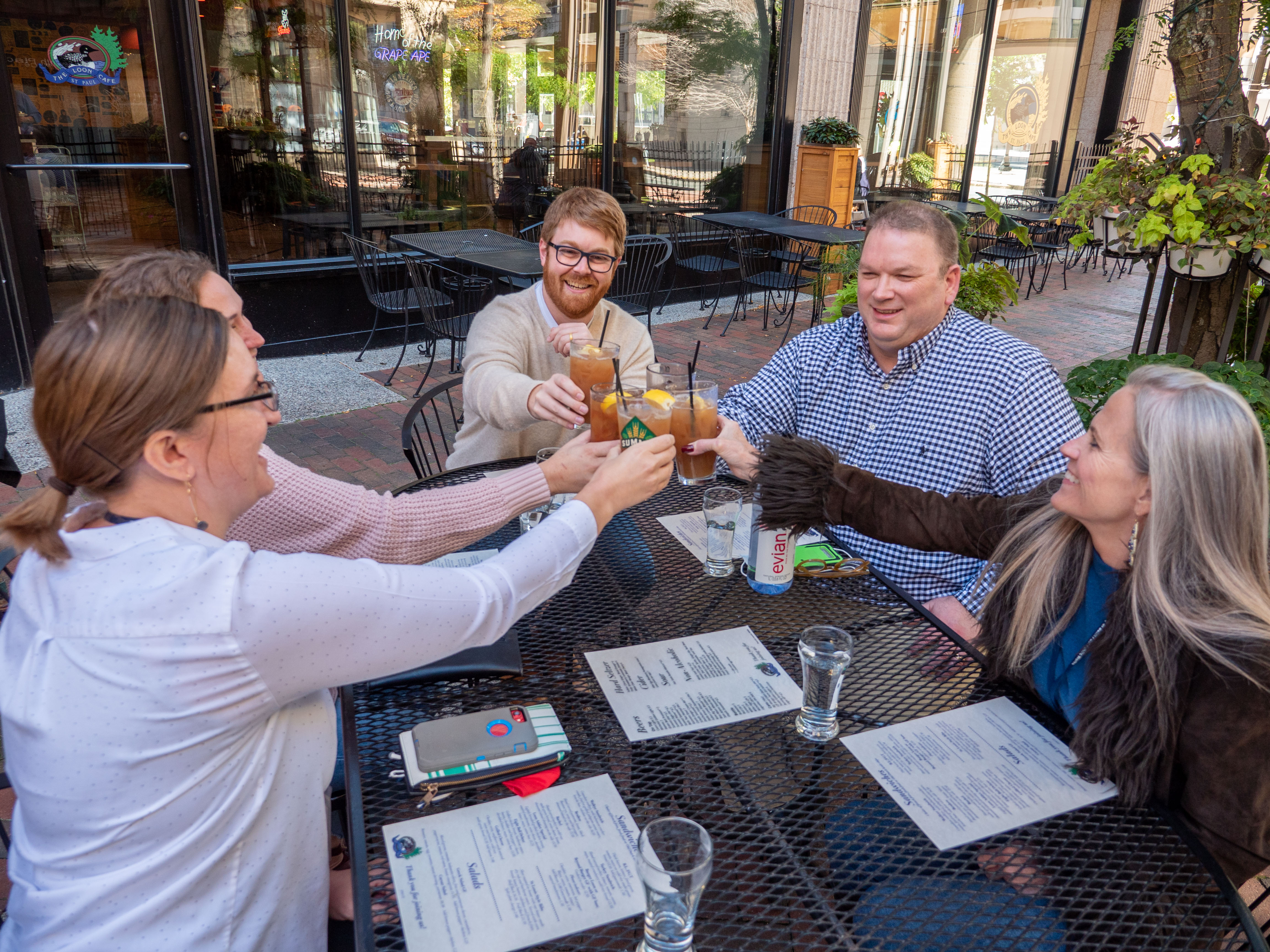 A group of people cheers with drinks, sitting at an outdoor table a patio alongside a restaurant.