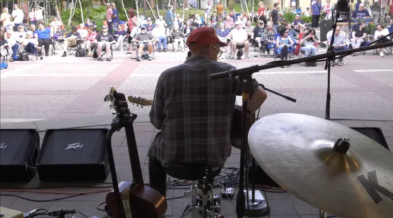 A musician plays a lunchtime concert at Mears Park on June 29, 2017. Photo of a man playing a guitar on an outdoor stage, facing an audience of people, sitting in chairs or walking around.