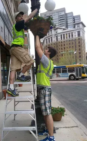 A person supports a hanging flower basket from below while another person on a ladder attaches the basket to a lamppost