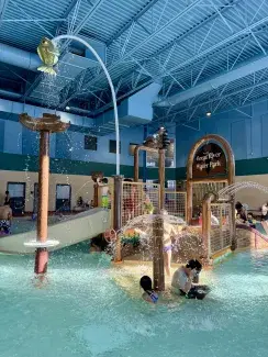 Great River Water Park - Children Pool Young children play in a shallow indoor pool. There are several water features, including multiple fountains, and a play structure.