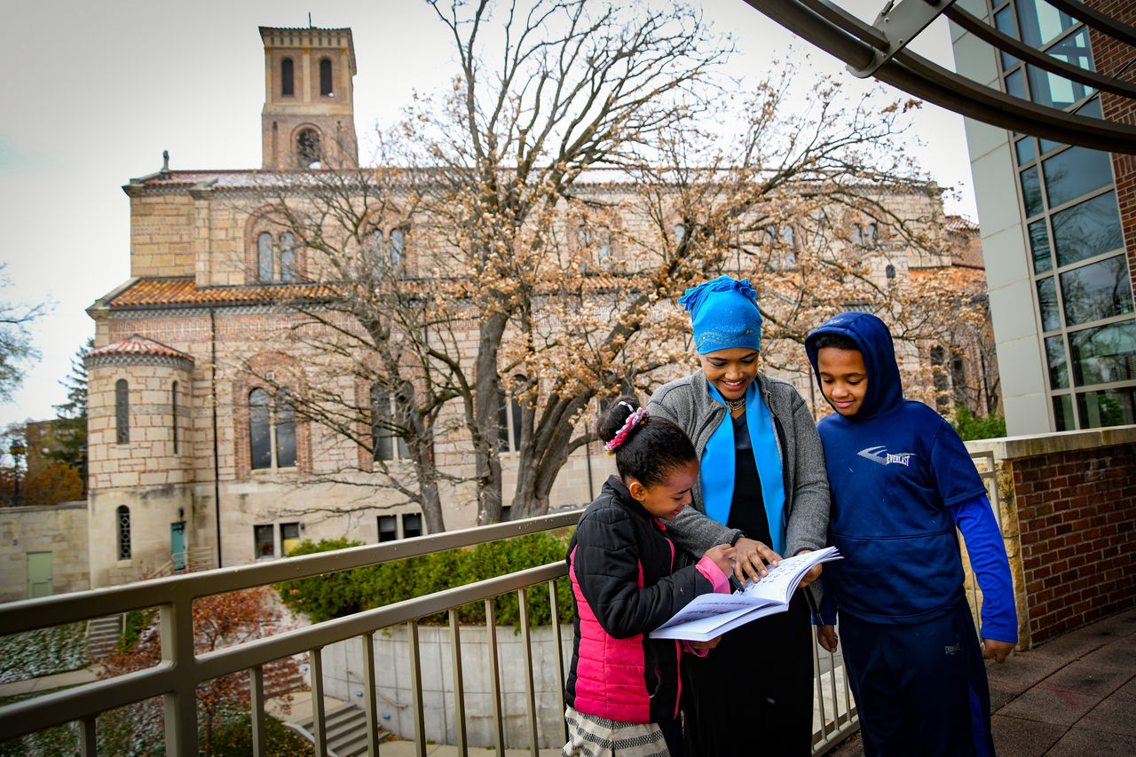 A woman with two school age children smile while reading a book outside on a Saint Paul college campus