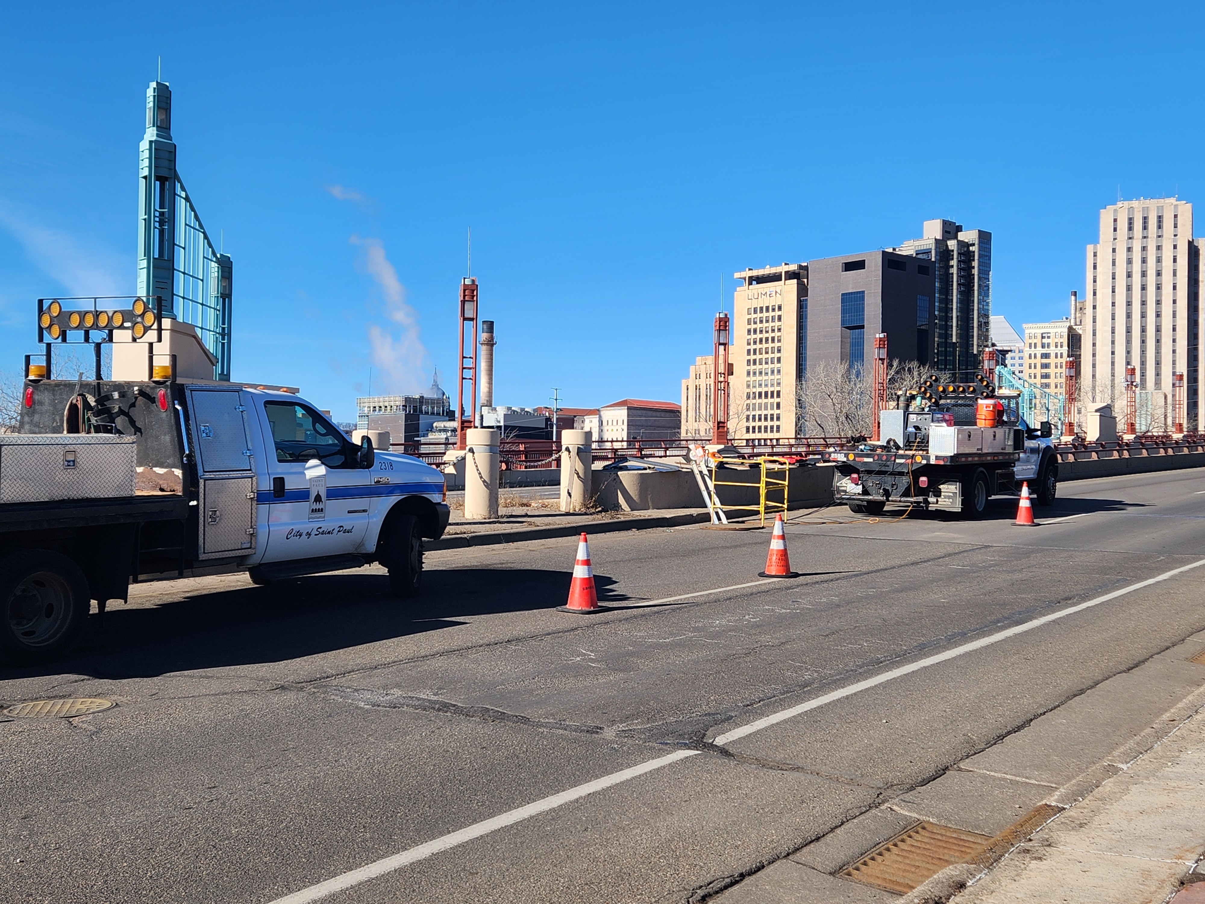 Two utility trucks from the City of Saint Paul are parked on a bridge, surrounded by orange traffic cones, with downtown skyscrapers visible in the background.