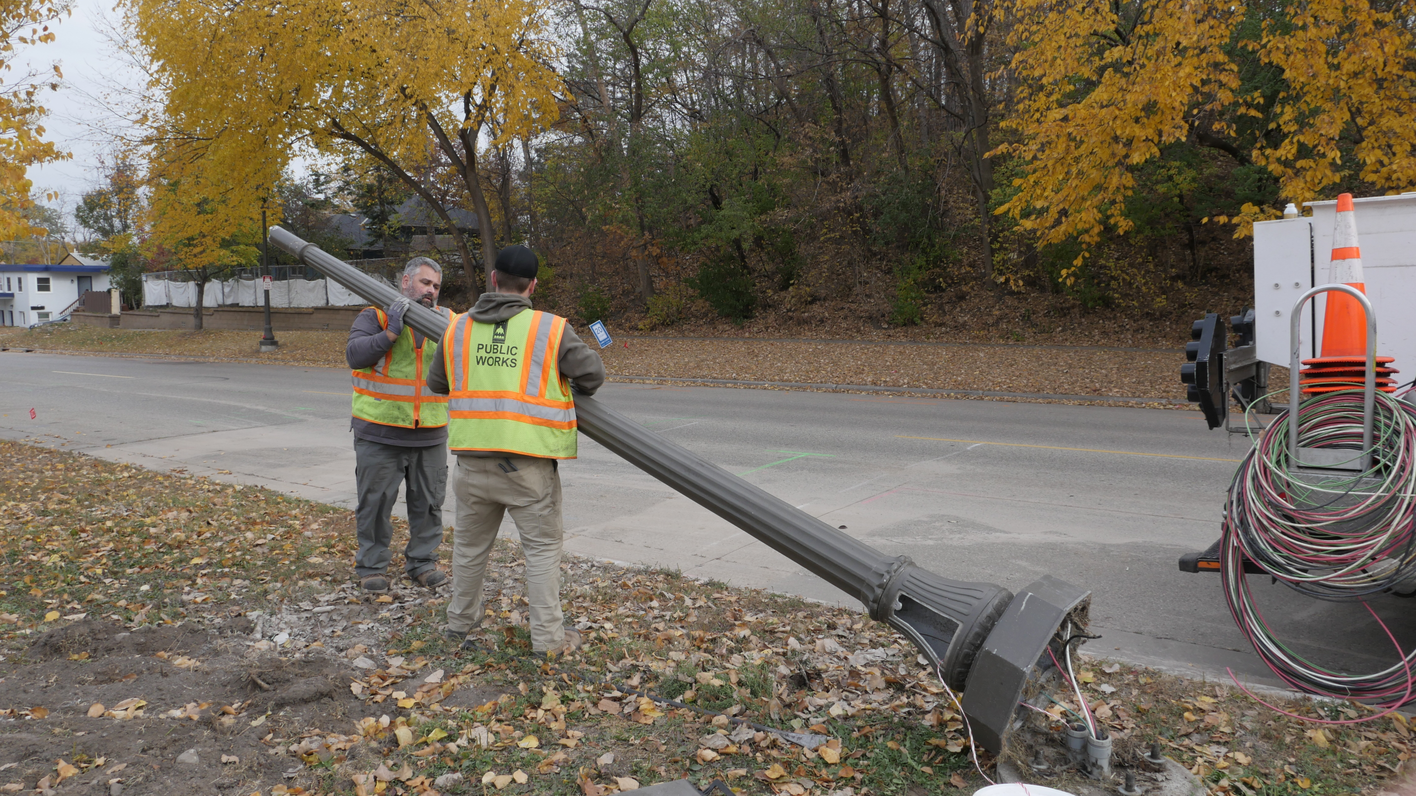 Two Public Works employees wearing high-visibility vests are removing a damaged streetlight pole on the side of a road lined with autumn trees. 