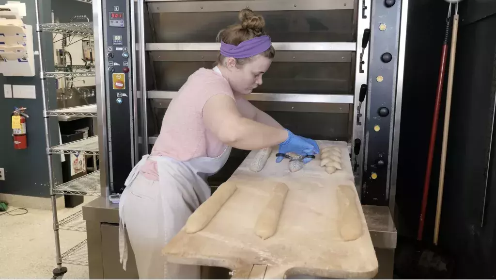 A young woman wearing a white apron, purple headband and disposable blue gloves scores loaves of bread before placing them in a commercial oven at a bakery.