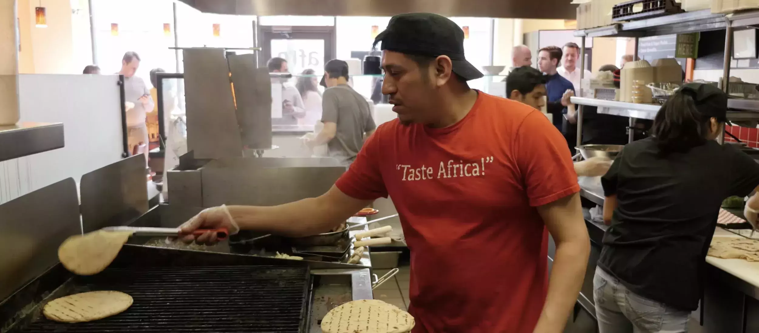 Employees in a restaurant kitchen work grilling bread and preparing a salad.