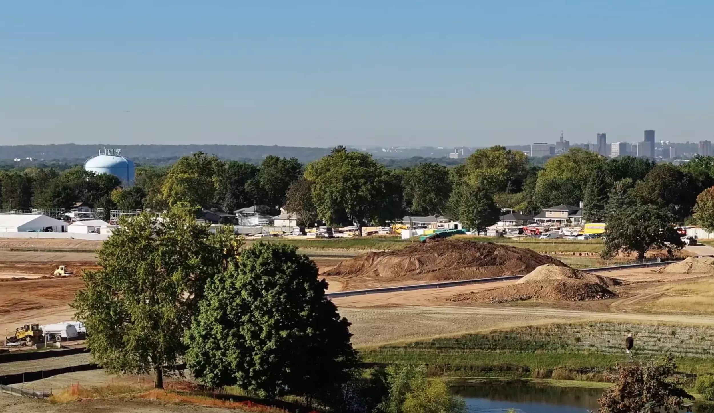 An aerial view of a large construction site with trees and bald eagle in foreground, city skyline visible in the distance.