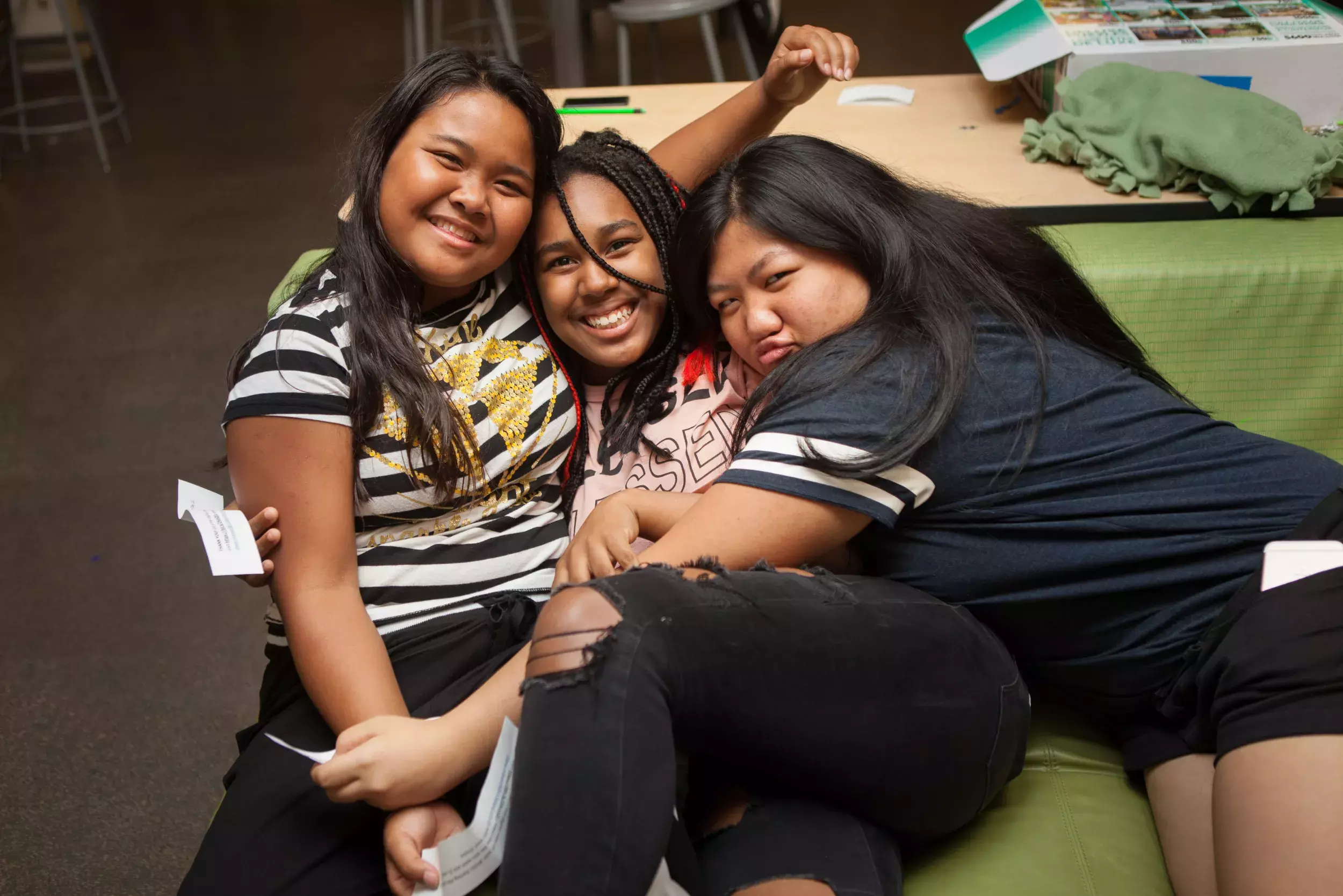 A diverse trio of young teenagers hugs on the couch while smiling. 