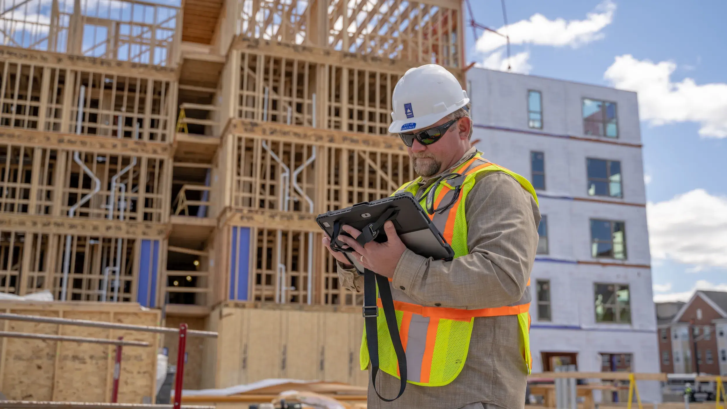 Man in City of Saint Paul construction helmet and high visibility vest examines tablet in front of a large residential building under construction.