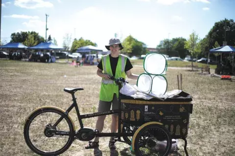 A person stands beside a cargo bike. The bike has bug nets and bug viewing boxes. There are canopy tents in the background.